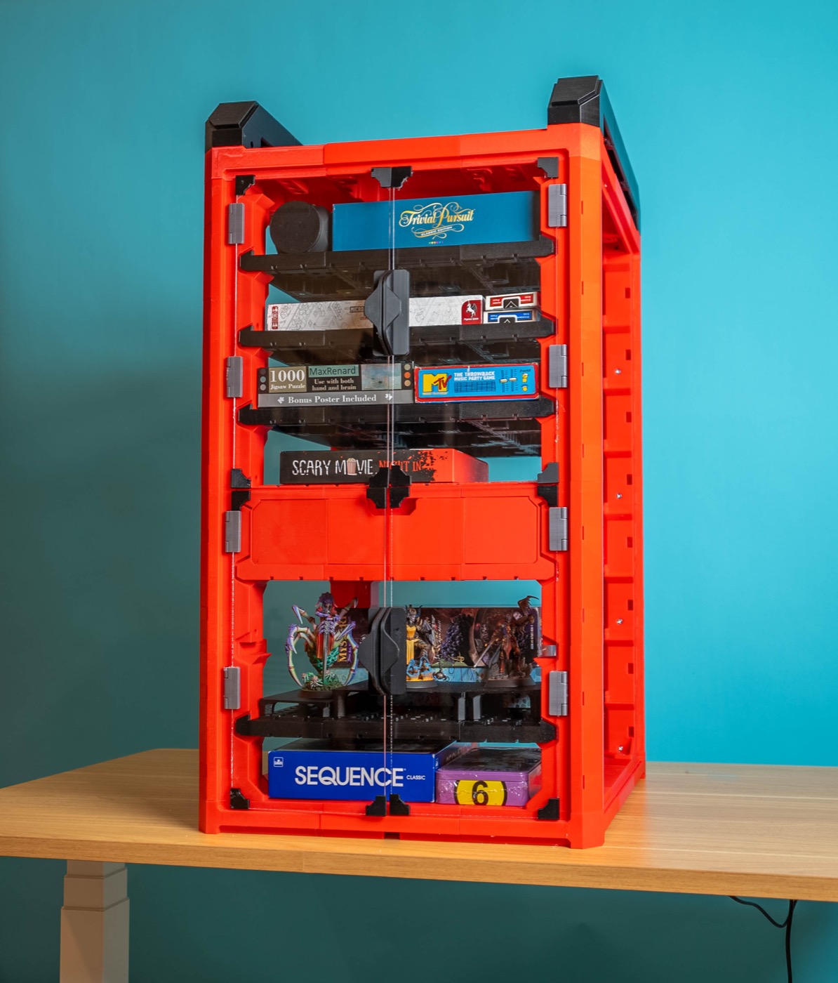 A red 3D-printed modular game crate on a desk, holding board games and painted miniatures